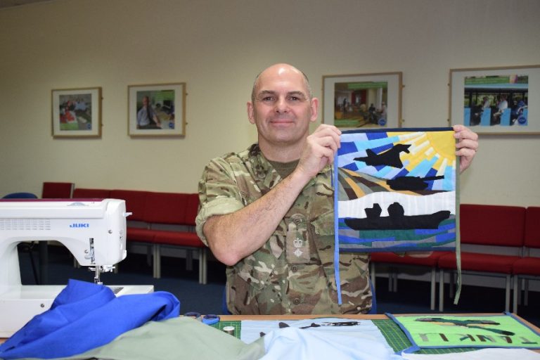A person in military uniform holds a fabric panel depicting flying birds and landscape artwork, seated at a table with a sewing machine and fabric pieces, creating Flags of Thanks.