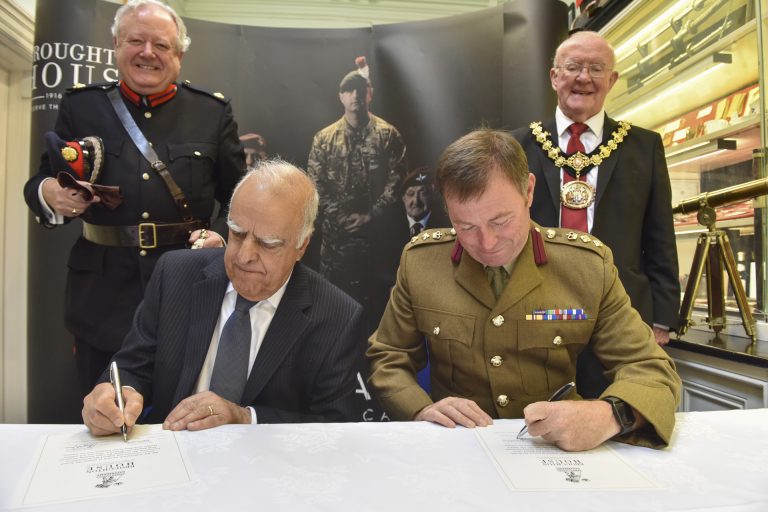 A veteran military officer and a civilian in suits sign documents at a table while two other men in official uniforms stand behind them, smiling.