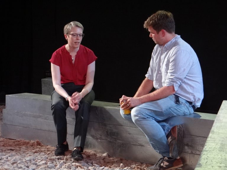 Two people sit on a stone bench, engaged in conversation. One wears a red blouse and black pants; the other wears a light blue shirt and jeans, holding a drink. The rocky ground surrounding them adds to the serene picture of civilian life on Civvy Street.