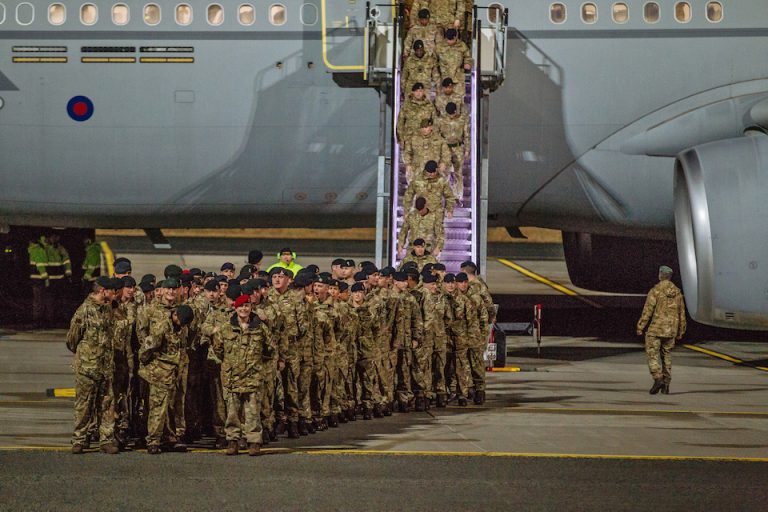 A group of soldiers in uniform disembark from a military aircraft, forming lines as they exit down a staircase onto the tarmac, ready to transition back into civilian life.