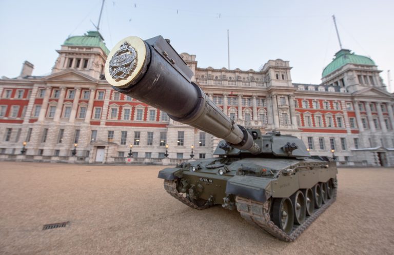 A military tank with a large cannon is displayed in front of a historic building with domed rooftops and multiple windows, symbolizing the transition from military to civilian life.