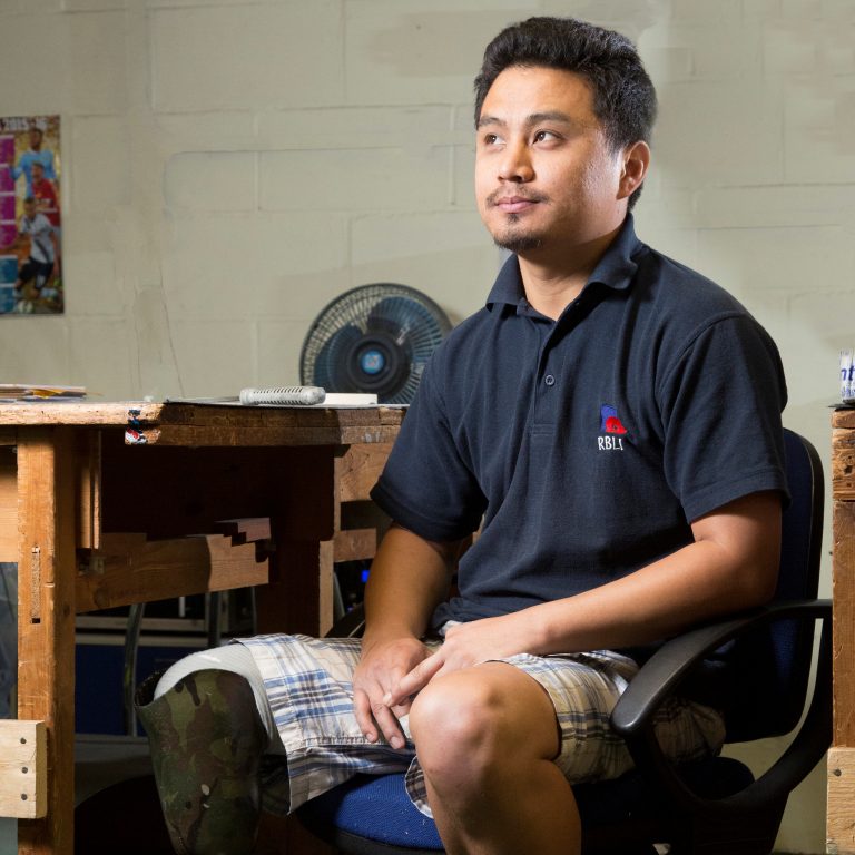 A veteran with a prosthetic leg sits on a chair beside a wooden table, wearing a black polo shirt and checkered shorts. Embracing life on Civvy Street, he enjoys the breeze of the fan visible in the background.