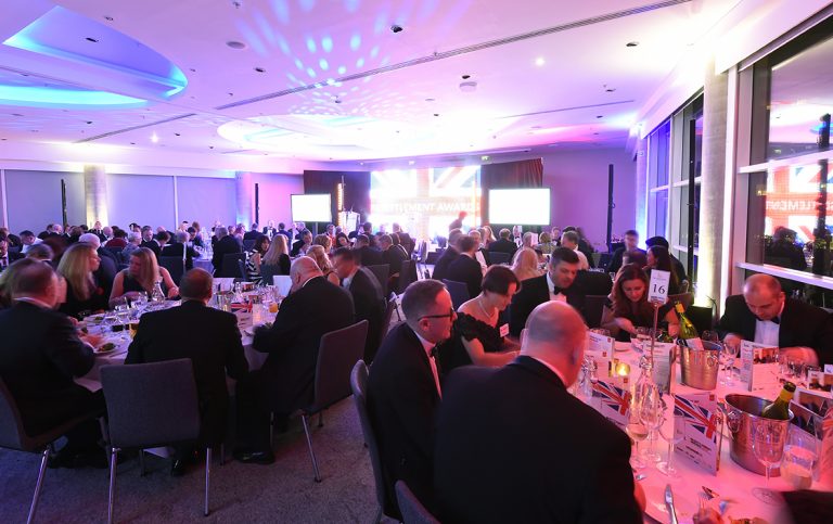 People in formal attire seated at round tables in a brightly lit banquet hall, attending an event with large screens displaying "AWARDS" in the background, celebrating outstanding achievements in resettlement and transitioning to civilian life.