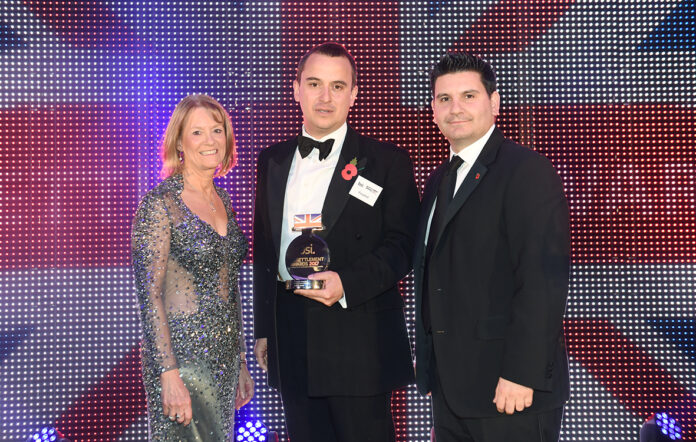 Three people stand in formal attire, with the man in the center holding an award. They are posed in front of a backdrop featuring the UK flag, celebrating their transition from military to Civvy Street.