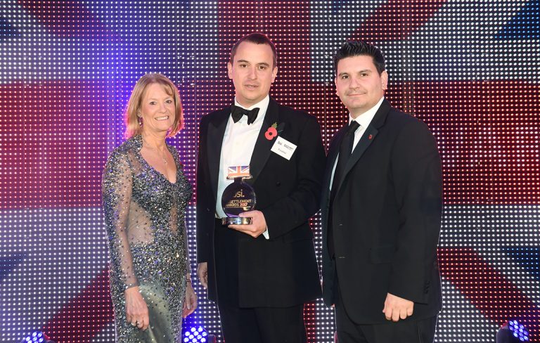 Three people stand in formal attire, with the man in the center holding an award. They are posed in front of a backdrop featuring the UK flag, celebrating their transition from military to Civvy Street.