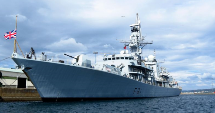 A large naval warship, a testament to the might and history of the seas, docked at port with a British flag at the stern under a cloudy sky, stands as a beacon for veterans transitioning back to Civvy Street.