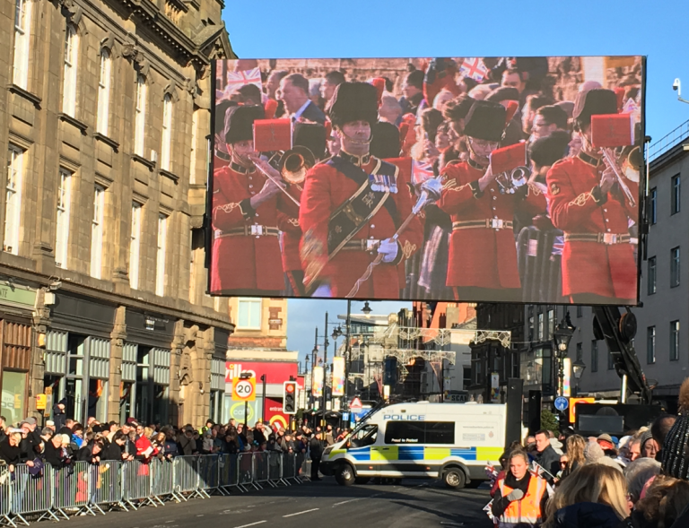 A military band, showcasing proud veterans, is displayed on a large screen during a bustling street parade. Crowds line the street, with a police vehicle maintaining order.