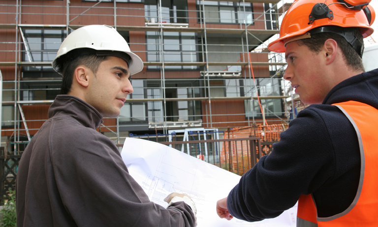 Two construction workers, one of whom is a veteran, wear helmets and safety gear as they discuss a set of blueprints at a construction site with scaffolding and a partially built structure in the background.