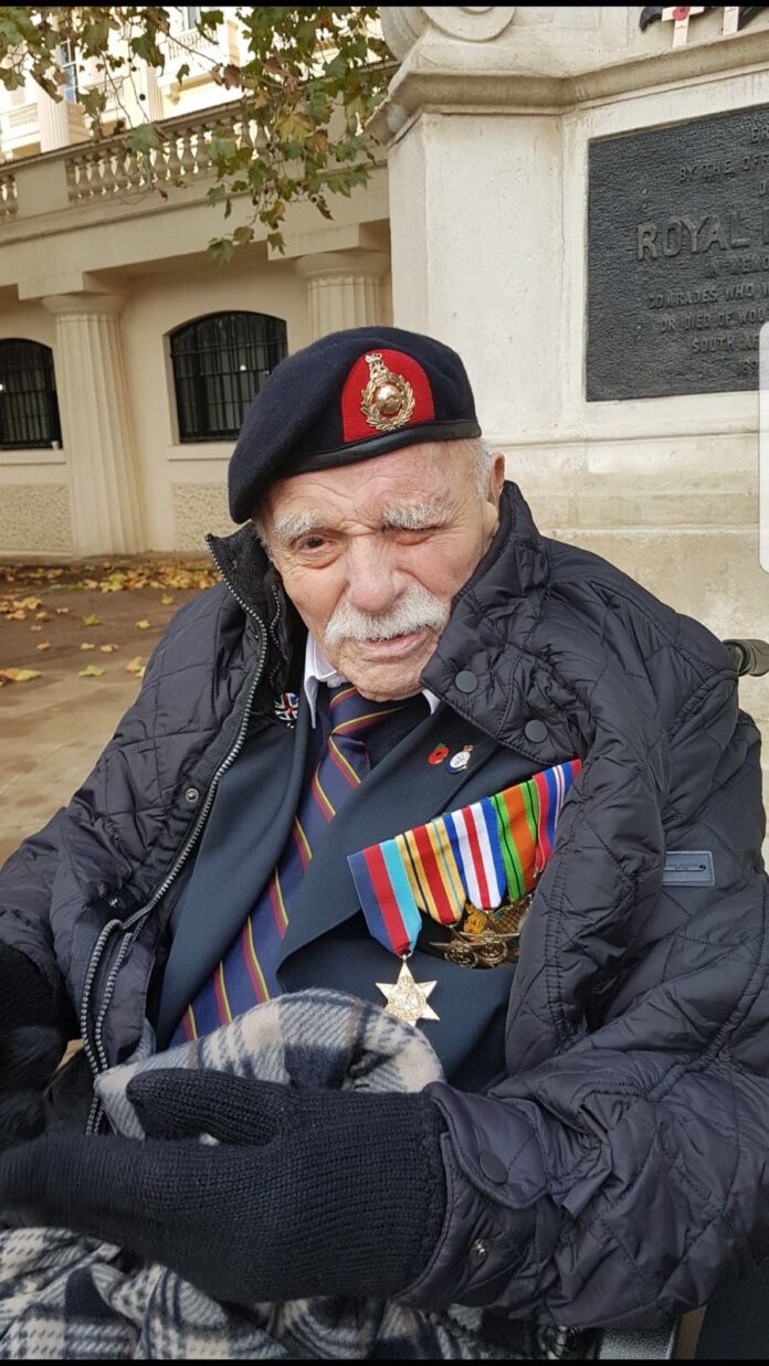 An elderly man wearing a military beret, various medals, and a striped tie sits outdoors against a stone monument, wrapped in a blanket and dressed warmly—a poignant reminder of the intersection between military service and civilian life.