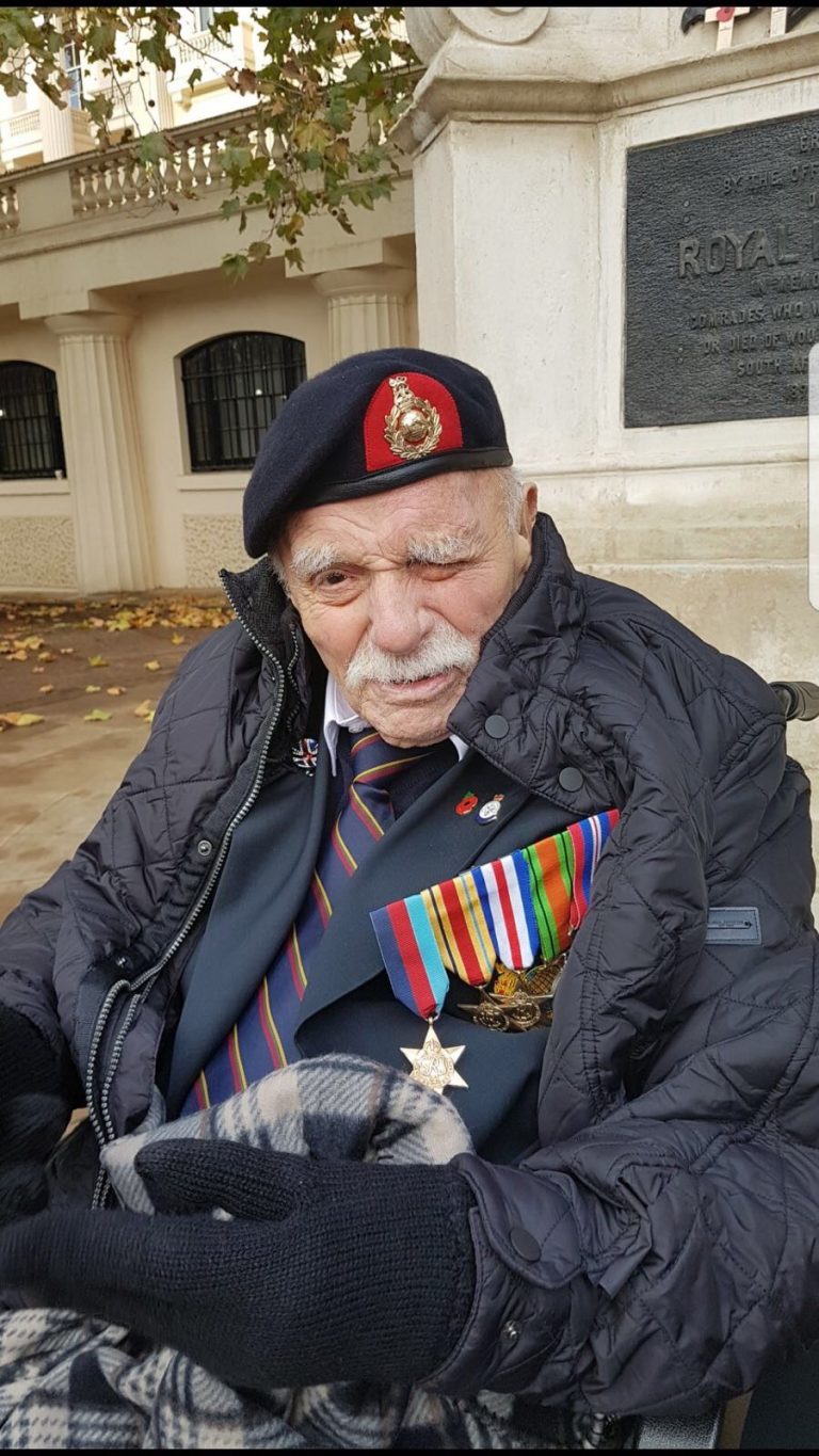 An elderly man wearing a military beret, various medals, and a striped tie sits outdoors against a stone monument, wrapped in a blanket and dressed warmly—a poignant reminder of the intersection between military service and civilian life.