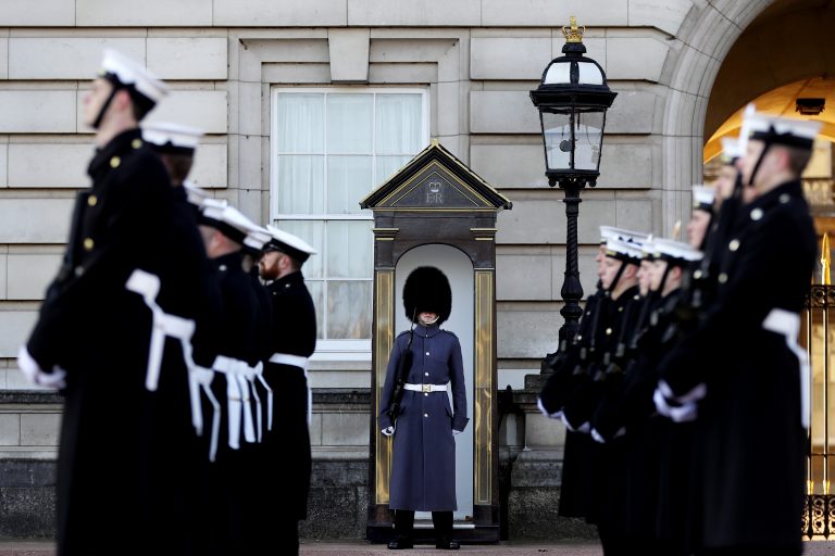 A veteran guard stands at attention under a sentry box, surrounded by a line of uniformed soldiers in a formal setting, reflecting on his journey from civvy street to this disciplined life.