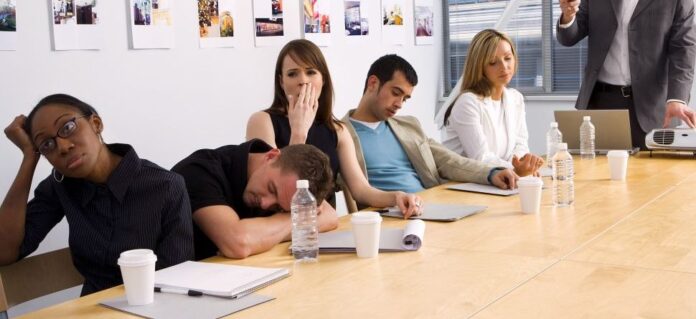 A group of people appear bored and tired in a meeting room, some yawning and some resting on the table, surrounded by paper cups, water bottles, and documents—a snapshot of civilian life that sharply contrasts with the discipline of military routine.