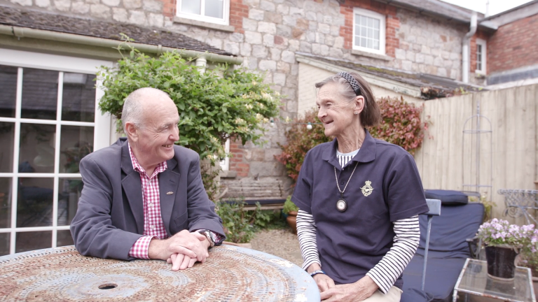 An elderly man and woman sit at a rustic outdoor table, smiling at each other in a garden setting, reminiscing about their resettlement journey from veteran life to Civvy Street. A stone building is visible in the background.