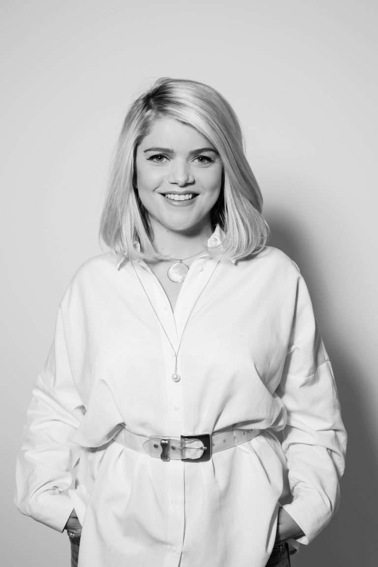 A person with shoulder-length hair, wearing a white shirt and belt, poses smiling with hands in pockets against a plain background, capturing the confidence of someone transitioning smoothly from veteran life to Civvy Street.