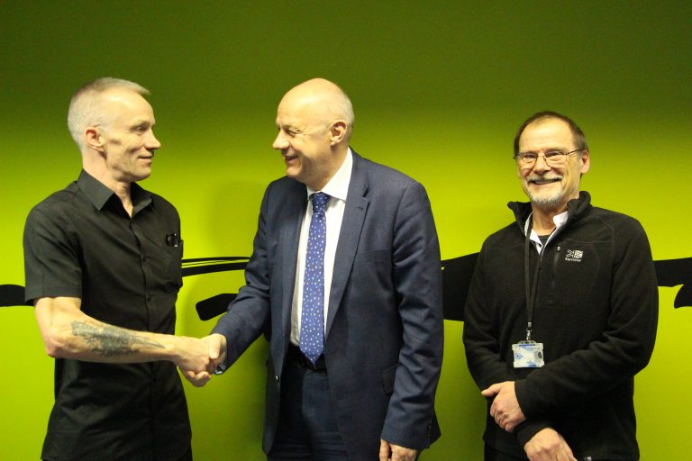 Three men stand in front of a green background. The man on the left, a veteran, and the man in the center are shaking hands. The man on the right is smiling, looking at the camera, symbolizing new beginnings in Civvy Street.