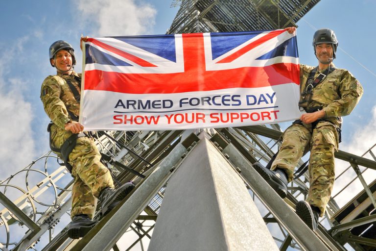 Two veteran soldiers in camouflage uniforms hold an "Armed Forces Day Show Your Support" flag, standing on a metal structure against the clear blue sky, proudly bridging their service and their journey back to Civvy Street.