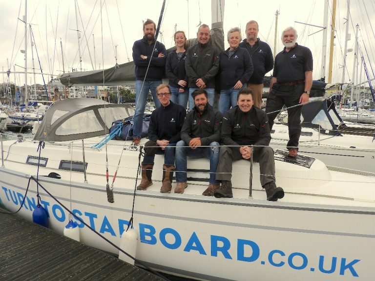 A group of nine veterans poses on a sailboat docked at a marina, with other boats in the background. The boat's side reads "Turn to Starboard.co.uk," symbolizing their journey from service life to Civvy Street.