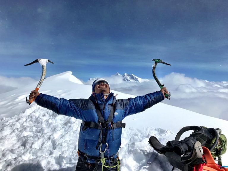 A veteran climber in a blue jacket stands triumphantly on a snowy peak, holding two ice axes with arms outstretched, surrounded by clouds and a mountain backdrop, embracing the challenge of transitioning from Civvy Street to conquer nature's heights.