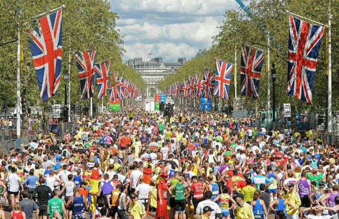 A large crowd of marathon runners on a wide street lined with Union Jack flags, surrounded by spectators and trees, mirrors the journey from military life to civvy street, where resettlement becomes a new race for many veterans.