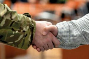 A veteran in military camouflage shakes hands with a person in a checkered shirt indoors, marking the beginning of their journey into civvy street.