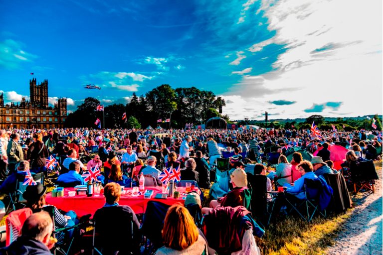 A large crowd gathers outdoors, with many people seated and holding British flags, celebrating a veteran's successful resettlement to civvy street. A historic building and trees are visible in the background under a partly cloudy sky.