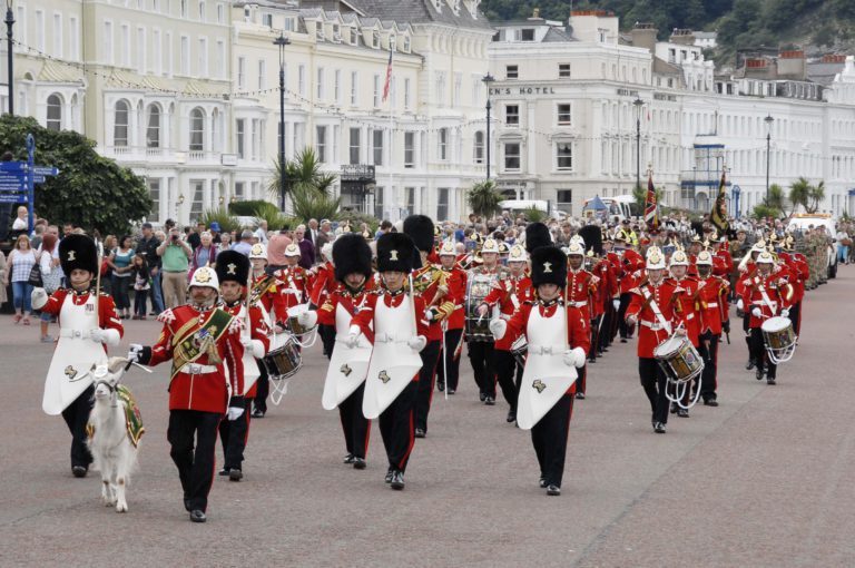 A marching band in red uniforms parades down Civvy Street, lined with spectators, featuring musicians with brass instruments and drums as if celebrating a veteran’s resettlement journey.