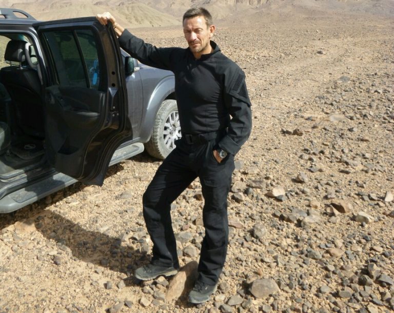 A veteran stands beside an open car door in a rocky desert landscape, wearing black clothing and boots, contemplating the vastness of life beyond Civvy Street.