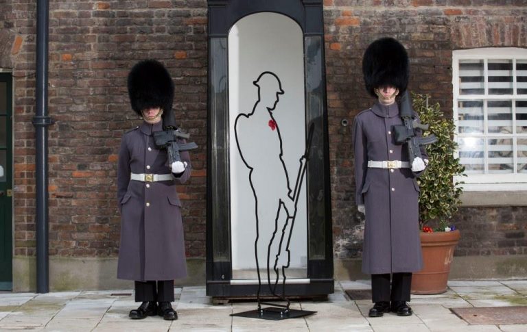 Two guards in traditional uniforms with tall hats stand beside a silhouette statue of a veteran soldier in front of a brick building.