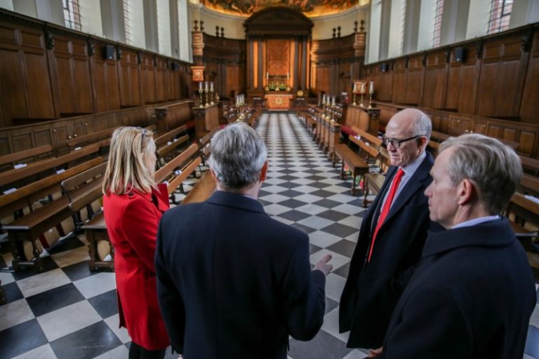 Four people, including a veteran adjusting to civvy street, stand in a church with checkered flooring, engaged in conversation. The church's wooden pews and altar provide a serene backdrop for their discussion.
