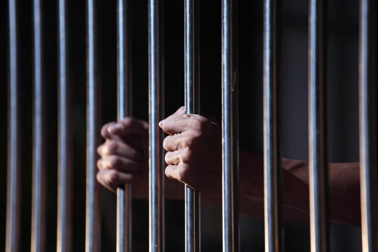 Hands gripping prison cell bars tightly in dim lighting, the veteran yearns for resettlement to Civvy Street.