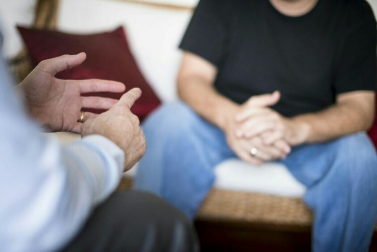 Two people seated and conversing, one gesturing with hands, the other sitting with hands clasped. In this casual setting, reminiscent of a veteran's transition to Civvy Street, cushions add a touch of comfort as they delve into the complexities of resettlement.