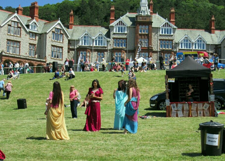 People in colorful attire gather on a grassy area in front of large, historic buildings, reminiscent of Civvy Street. A black tent and a waste bin are visible, with more people and activities in the background, symbolizing the vibrancy of resettlement.