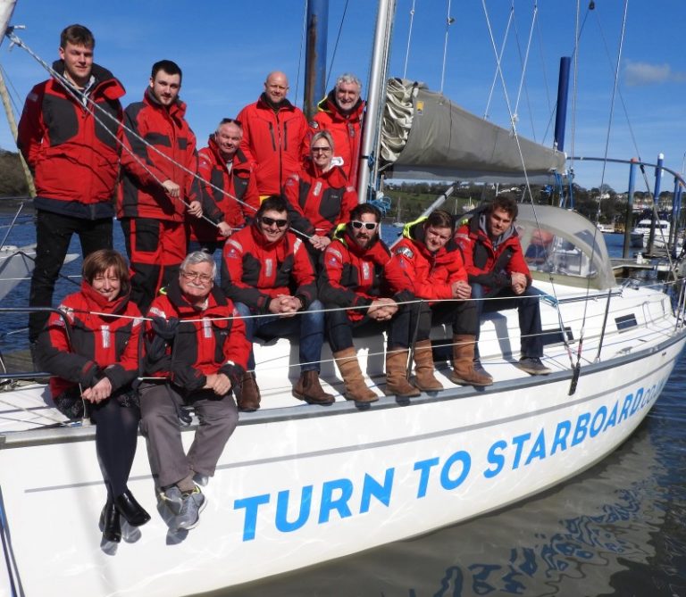 A group of veterans in red jackets sits on the sailboat "Turn to Starboard," docked at a marina, enjoying their newfound camaraderie as they adjust to Civvy Street.