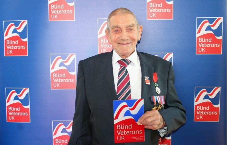 An elderly veteran in a suit holds a plaque with the Blind Veterans UK logo, standing against a backdrop with the same logo, symbolizing his journey from Civvy Street to successful resettlement.
