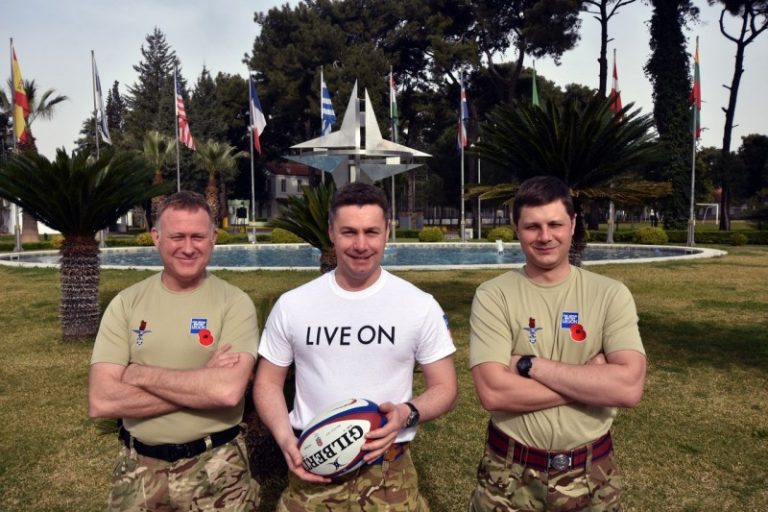 Three veterans in military uniform stand on grass, with one holding a rugby ball. Behind them is a fountain and several international flags, symbolizing their journey from service to Civvy Street.