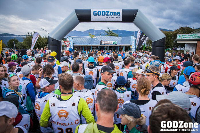 Veteran participants gather at the starting line of the GODZone adventure race in Fiordland, New Zealand, under an inflatable arch on a cloudy day.