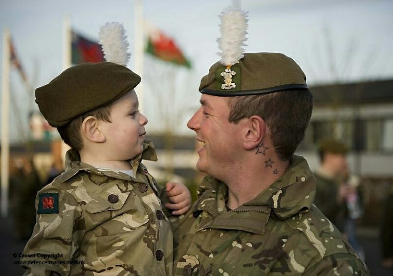 A veteran soldier in uniform holds a smiling child wearing a matching beret and uniform, symbolizing the transition from military life to civvy street. Flags are visible in the background.