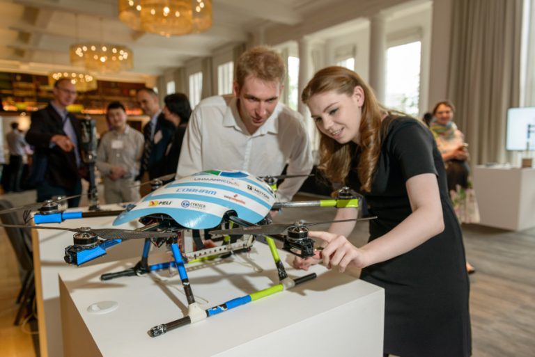 Two veterans examine a drone on display in a room, navigating their resettlement into Civvy Street, with a few intrigued onlookers in the background.