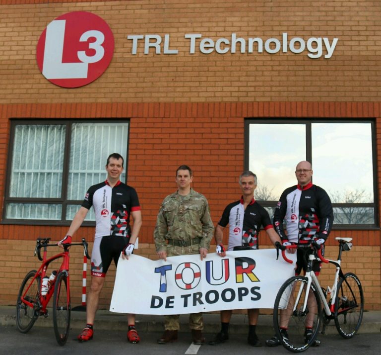 Four people holding a "Tour de Troops" banner stand proudly in front of a building with "L3 TRL Technology" signage. Three, dressed in cycling gear, are beside their bicycles, while one, a veteran adjusting to Civvy Street, stands tall in military uniform.