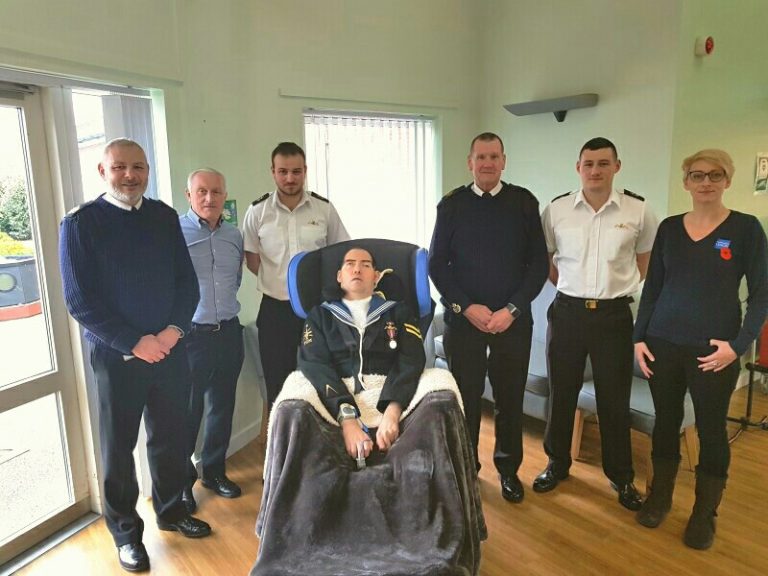 A group of seven people, including a veteran in a wheelchair, pose indoors by windows, some in uniform.