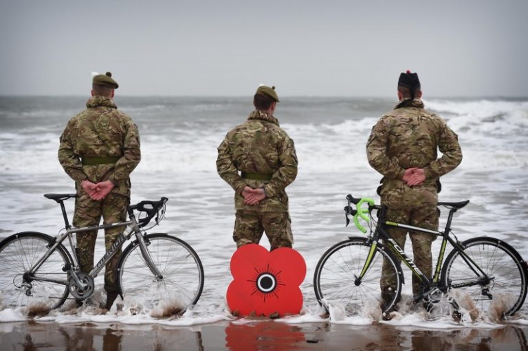 Three soldiers in military uniform stand on a beach with bicycles and a large red poppy emblem, reflecting on their journey from veteran to civvy street as they face the ocean.