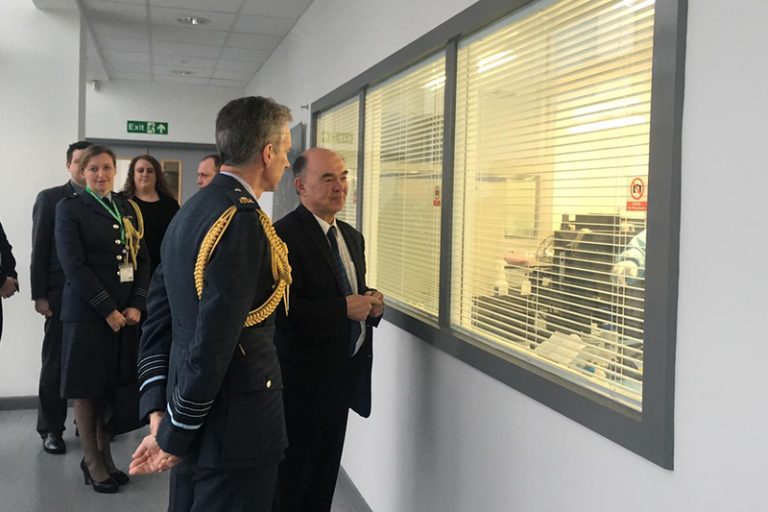 A group of individuals in formal attire stands in a corridor, observing through a window with blinds into a room, discussing a veteran's transition to Civvy Street.