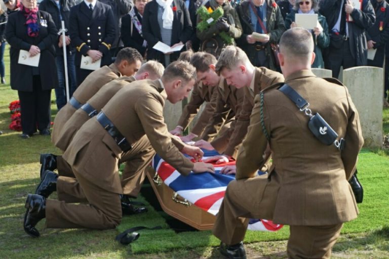 Soldiers in uniform kneel with reverence, placing a flag-draped coffin of a veteran into the grave during a ceremonial service, as a crowd of onlookers pays their respects in the background.