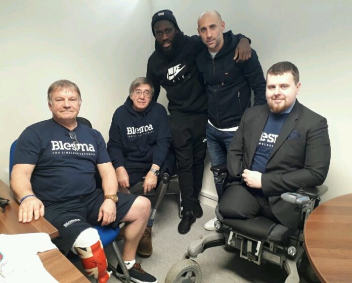 Five men in a room, three seated and two standing, casually gathered. Some sport Blesma clothing, signaling their veteran status. Among them, one sits in a wheelchair. The scene captures a moment of camaraderie as they discuss life on Civvy Street and resettlement challenges.