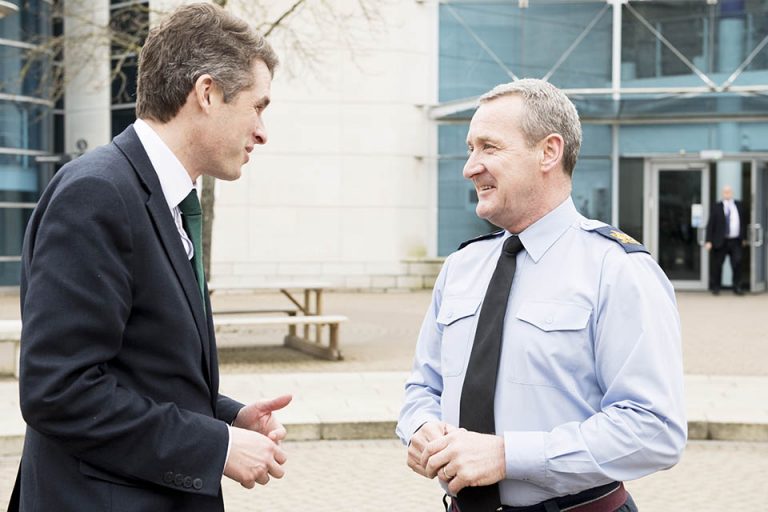 Two men in conversation outdoors, one in a suit and the other a veteran in a uniform, stand before a modern building.