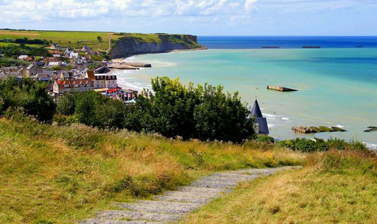 Coastal village with houses, green fields, and a cliff overlooking the sea. A path leads through grassy terrain towards the village and beach, embodying the spirit of resettlement for those transitioning to Civvy Street.