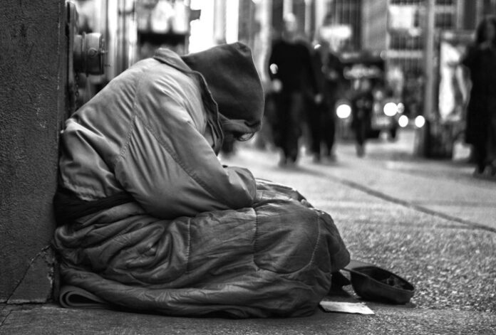A veteran, wrapped in layers of clothing with a hat covering their head, sits on the sidewalk. The urban street scene, characteristic of Civvy Street, buzzes around them with blurred figures and lights in the background.