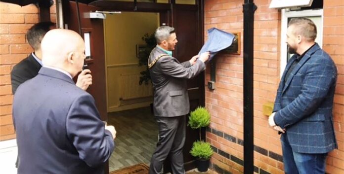 A veteran in a suit removes a cover from a plaque on a brick wall, observed by three other men, marking his transition from civvy street.
