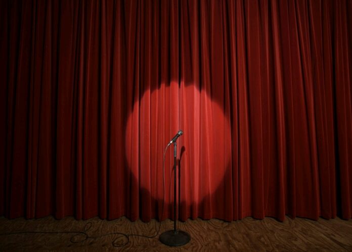 A microphone stands in a spotlight on a wooden stage, where stories of resettlement echo through closed red curtains.