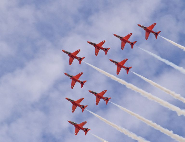 Nine red military jets fly in formation, trailing white smoke against a cloudy blue sky.